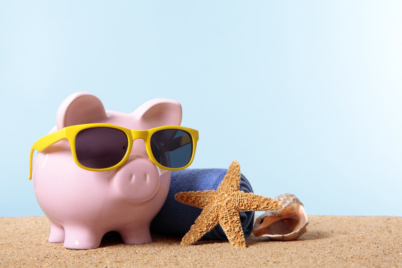 Pink piggy bank on a beach with sunglasses and beach towel. Space for copy.